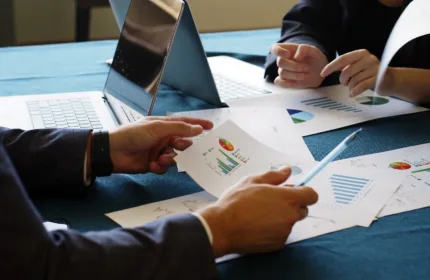 Two business professionals review workforce reports and charts at a table with open laptops.